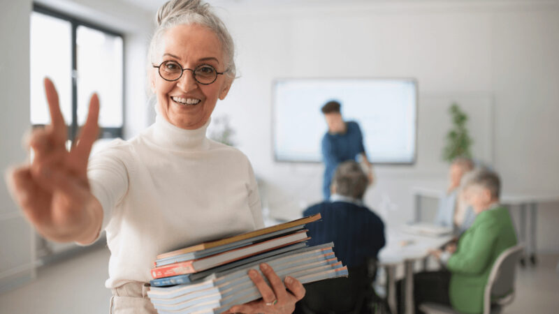 Eine ältere Dame, die nach der Teilnahme an einem Kurs an der Volkshochschule strahlt vor Freude. Sie zeigt das Victory-Zeichen als Ausdruck ihrer Begeisterung und des persönlichen Erfolgs.